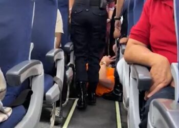 Un pasajero que se encuentra en el piso de un avión mientras un oficial de policía se encuentra sobre ellos.