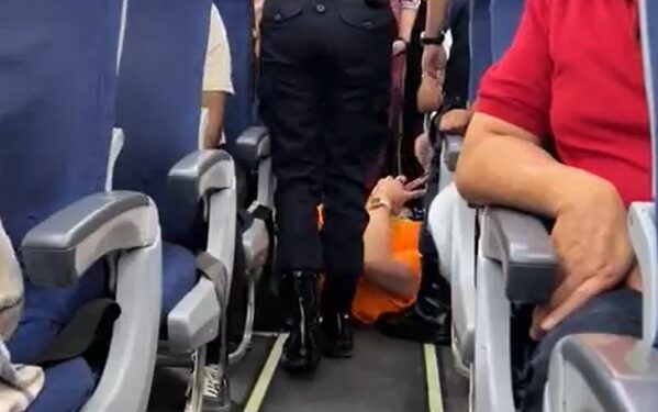 Un pasajero que se encuentra en el piso de un avión mientras un oficial de policía se encuentra sobre ellos.
