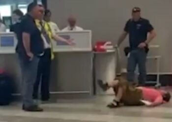 Persona acostada en el piso en un aeropuerto; Varias personas paradas cerca.