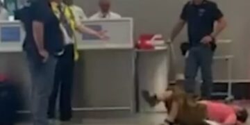 Persona acostada en el piso en un aeropuerto; Varias personas paradas cerca.