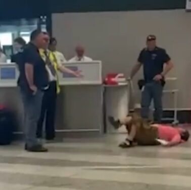 Persona acostada en el piso en un aeropuerto; Varias personas paradas cerca.