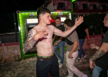 Un joven sin camisa con tatuajes gestos cerca de un cajero automático.