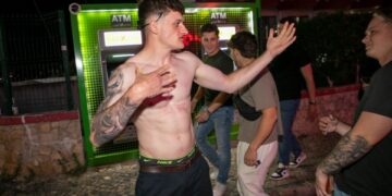 Un joven sin camisa con tatuajes gestos cerca de un cajero automático.