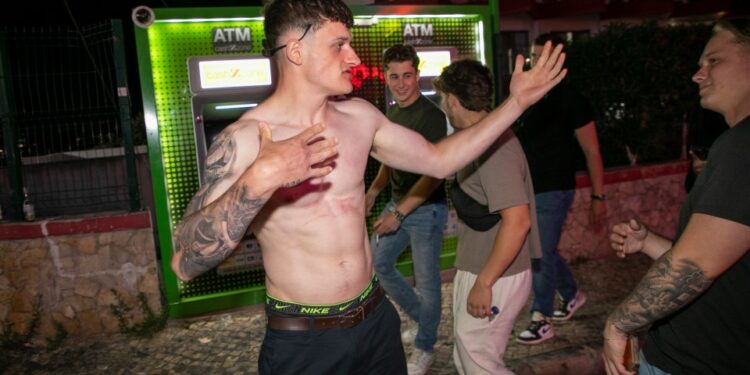 Un joven sin camisa con tatuajes gestos cerca de un cajero automático.