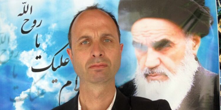 Un hombre con un traje posando con una gran foto de ayatolá Jomeini en el fondo.