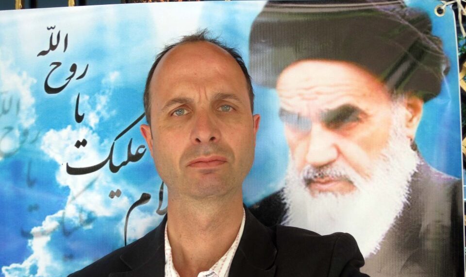 Un hombre con un traje posando con una gran foto de ayatolá Jomeini en el fondo.