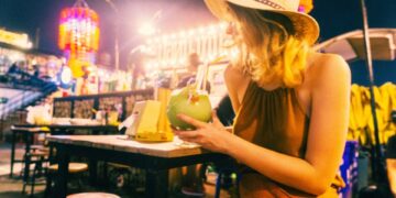 Mujer con sombrero de paja bebiendo un cóctel en un mercado nocturno.