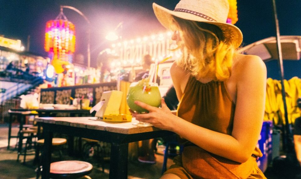 Mujer con sombrero de paja bebiendo un cóctel en un mercado nocturno.
