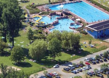 Vista aérea de un complejo de piscinas al aire libre con un tobogán de agua, rodeado por un área cubierta de hierba con gente tomada por el sol y un estacionamiento.