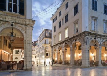 Plaza del pueblo al anochecer con cafés debajo de columnatas arqueadas.