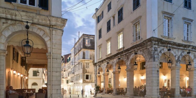 Plaza del pueblo al anochecer con cafés debajo de columnatas arqueadas.