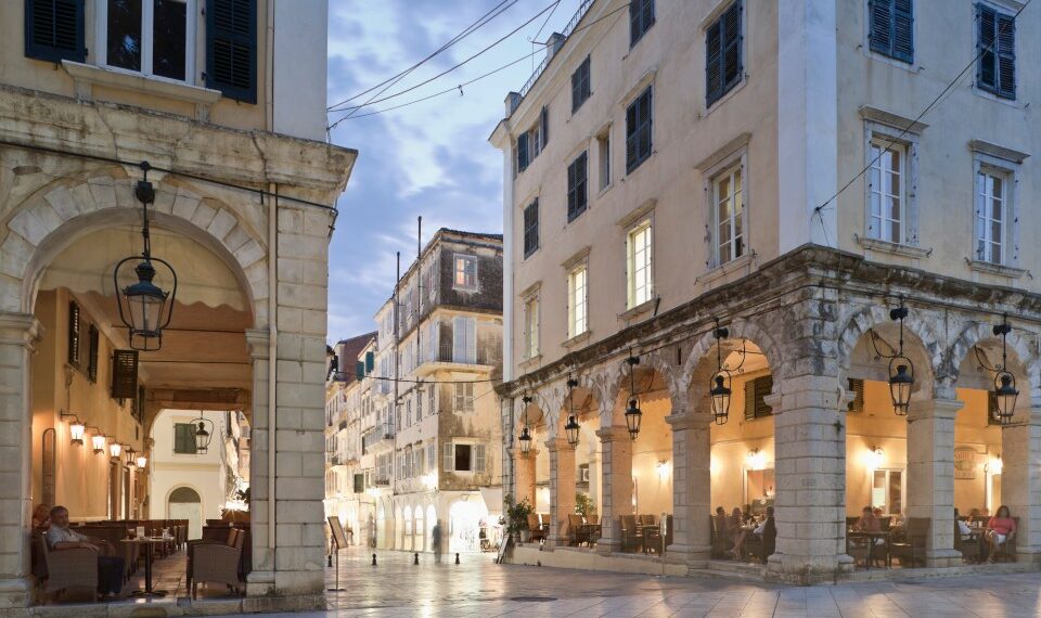 Plaza del pueblo al anochecer con cafés debajo de columnatas arqueadas.