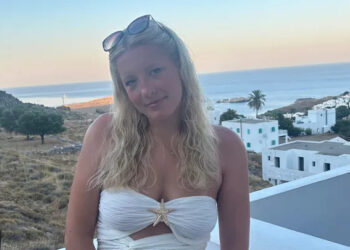 Una mujer joven con cabello rubio con un top blanco en un balcón con vistas al océano.