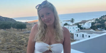 Una mujer joven con cabello rubio con un top blanco en un balcón con vistas al océano.