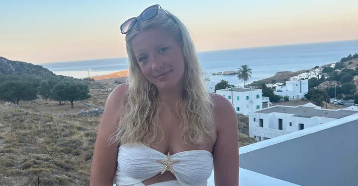 Una mujer joven con cabello rubio con un top blanco en un balcón con vistas al océano.