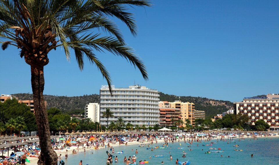 Playa llena de gente en Magaluf, Mallorca, con hoteles en el fondo.