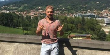 Hombre con camisa rosa y pantalones cortos azules claros que hacen un letrero Shaka frente a una ciudad de montaña.
