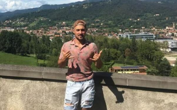 Hombre con camisa rosa y pantalones cortos azules claros que hacen un letrero Shaka frente a una ciudad de montaña.