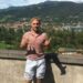 Hombre con camisa rosa y pantalones cortos azules claros que hacen un letrero Shaka frente a una ciudad de montaña.