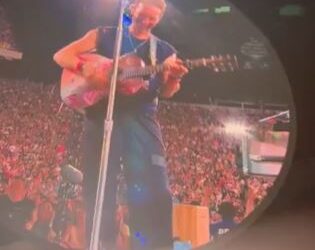 Chris Martin tocando la guitarra en una pantalla grande en un concierto.
