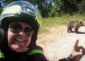 Un motociclista sonriendo y apuntando a un oso en una carretera.