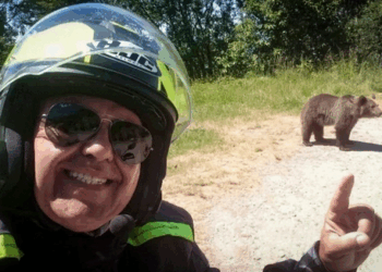 Un motociclista sonriendo y apuntando a un oso en una carretera.