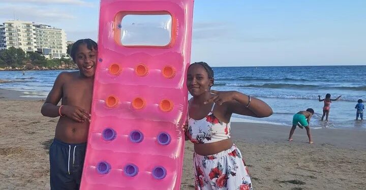 Dos niños en una playa con una balsa inflable rosa.