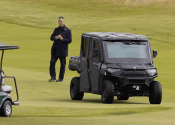 Hombre de pie en un campo de golf cerca de un carrito de golf y un vehículo utilitario negro.