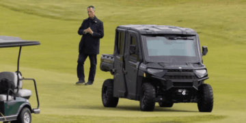 Hombre de pie en un campo de golf cerca de un carrito de golf y un vehículo utilitario negro.