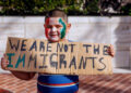 Un chico lttle con pintura de cara roja, blanca y verde contiene un letrero escrito a mano que lee "No tenemos los inmigrantes" mientras participa en una protesta al aire libre
