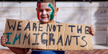 Un chico lttle con pintura de cara roja, blanca y verde contiene un letrero escrito a mano que lee "No tenemos los inmigrantes" mientras participa en una protesta al aire libre