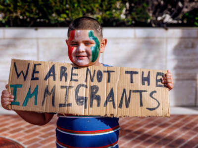 Un chico lttle con pintura de cara roja, blanca y verde contiene un letrero escrito a mano que lee "No tenemos los inmigrantes" mientras participa en una protesta al aire libre