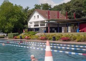 Piscina al aire libre con nadadores y un edificio en el fondo.