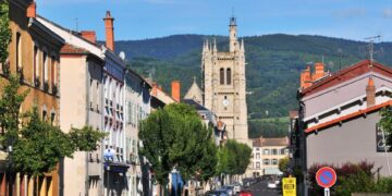Escena callejera en Ambert, Francia, que muestra edificios, autos y una torre de la iglesia en el fondo.