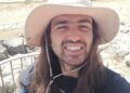 Selfie de un hombre sonriente con cabello largo con un sombrero de sol, tomado al aire libre.
