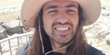 Selfie de un hombre sonriente con cabello largo con un sombrero de sol, tomado al aire libre.
