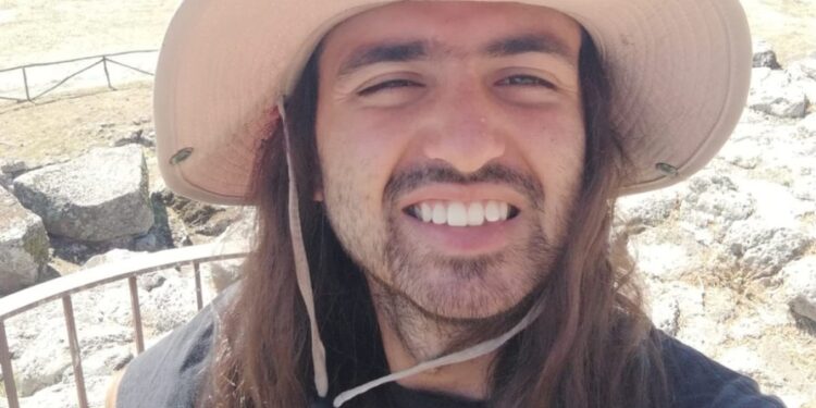 Hombre con cabello largo y un sombrero sonriendo en un paisaje.