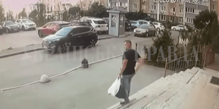 El hombre que lleva una bolsa sube escalones hacia un edificio con autos estacionados en el fondo.