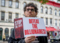 Un manifestante tiene un letrero que lee "Detener los cortes; Derrotar a los multimillonarios" mientras participa en una demostración al aire libre