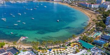 Vista aérea de la Bahía de San Antonio, Ibiza, que muestra una playa, botes y un resort con una piscina y muchos paraguas amarillos.