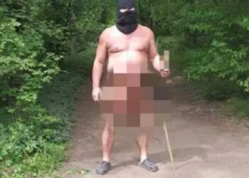 Un hombre desnudo que lleva un balaclava y las zuecos sostienen un juguete sexual en un palo en una zona boscosa.