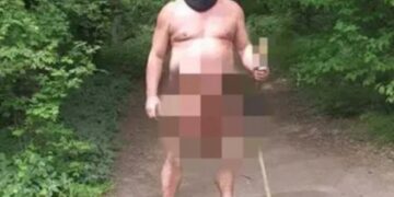 Un hombre desnudo que lleva un balaclava y las zuecos sostienen un juguete sexual en un palo en una zona boscosa.