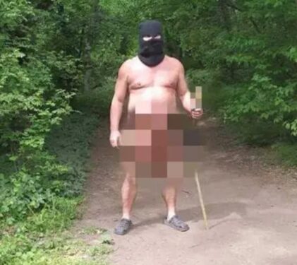 Un hombre desnudo que lleva un balaclava y las zuecos sostienen un juguete sexual en un palo en una zona boscosa.