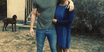 Hombre y mujer que abrazan al aire libre con un perro en el fondo.