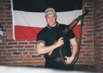 Foto de un joven sosteniendo un rifle, parado frente a una bandera alemana.