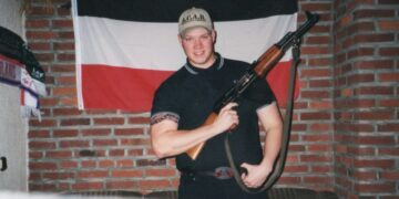 Foto de un joven sosteniendo un rifle, parado frente a una bandera alemana.