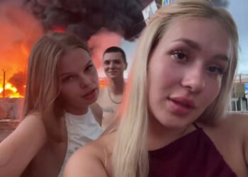 Dos mujeres jóvenes se filman frente a un depósito de aceite en llamas.