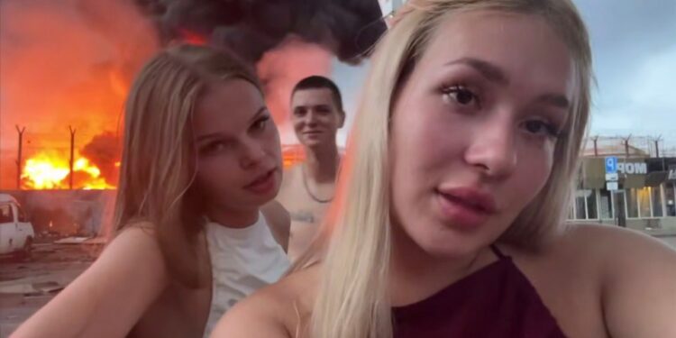 Dos mujeres jóvenes se filman frente a un depósito de aceite en llamas.