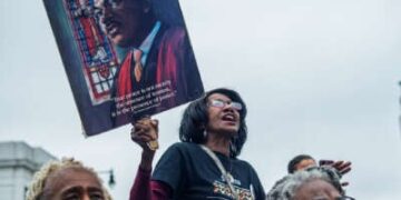 Una mujer canta junto a otros manifestantes mientras sostiene un letrero que descubre la cara del reverendo del reverendo Martin Luther King
