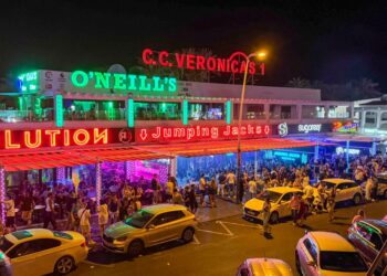 Escena de la calle nocturna con bares llenos de gente y autos estacionados.